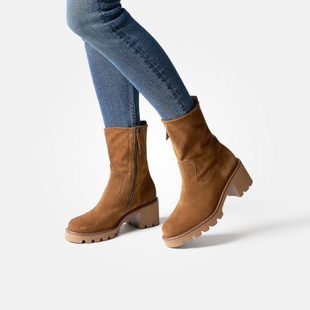 A lady wearing brown Paul Green Wanda ankle boots paired with blue jeans, set against a light gray background.